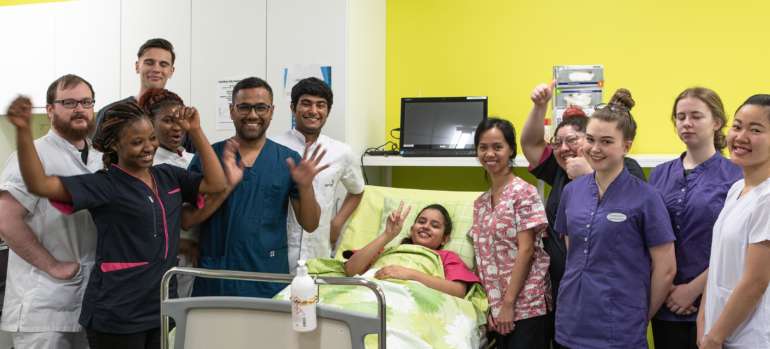 Henkilöitä eri taustoista ryhmäkuvassa sairaalasängyn ympärillä hoitajan työvaatteissa ja katsomassa iloisesti kameraan.